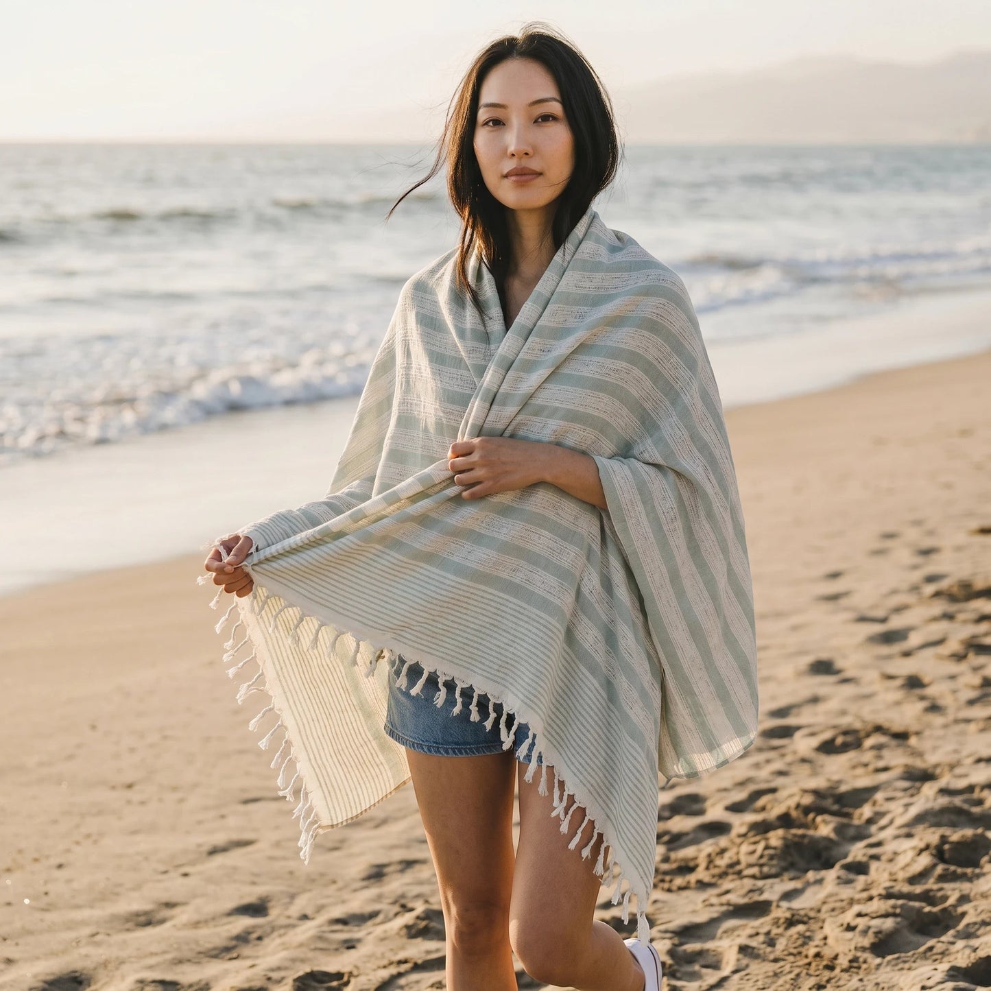 Naqada Hand Crafts handwoven cotton shawl with sage green stripes and fringe, worn by a woman on a sandy beach.