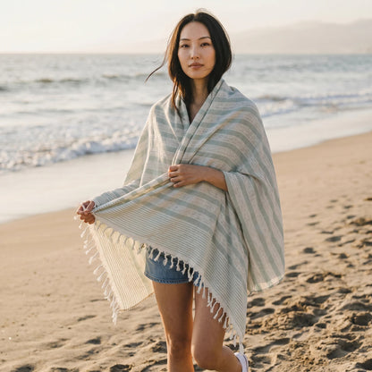 Naqada Hand Crafts handwoven cotton shawl with sage green stripes and fringe, worn by a woman on a sandy beach.